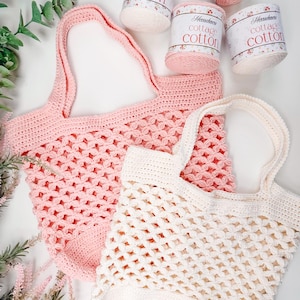 Two crocheted tote bags, one pink and one white, are displayed on a white surface beside four skeins of cotton yarn and sprigs of greenery—perfect as a handmade gift for fans of vintage clothes.
