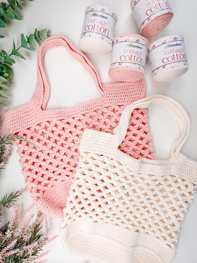 Two crocheted tote bags, one pink and one white, are displayed on a white surface beside four skeins of cotton yarn and sprigs of greenery—perfect as a handmade gift for fans of vintage clothes.