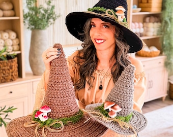 May include: Three crocheted witch hats in black, brown, and gray. Each hat features a wide brim and a pointed crown, decorated with green moss, natural twine, and small artificial mushrooms attached to the base of the crown.