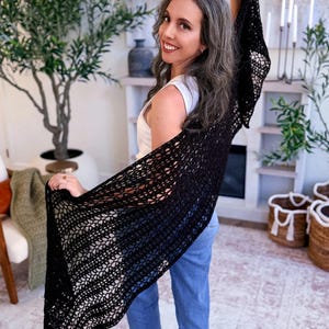 A woman with long gray hair, wearing a white top and blue jeans, holds up a large black crocheted shawl indoors—a perfect handmade gift—surrounded by plants and baskets in the background.