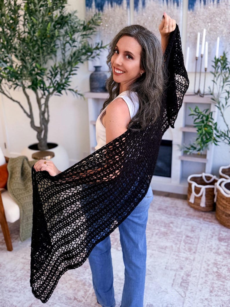 A woman with long gray hair, wearing a white top and blue jeans, holds up a large black crocheted shawl indoors—a perfect handmade gift—surrounded by plants and baskets in the background.