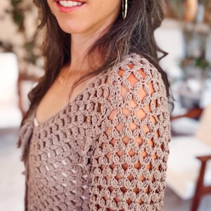 May include: A long-sleeved, light brown crochet top featuring an intricate, open-work diamond pattern throughout the fabric. The garment has a V-neckline and a fitted silhouette, showcasing the detailed texture of the yarn and the repeating stitch design.
