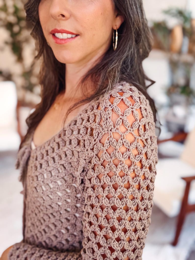 May include: A long-sleeved, light brown crochet top featuring an intricate, open-work diamond pattern throughout the fabric. The garment has a V-neckline and a fitted silhouette, showcasing the detailed texture of the yarn and the repeating stitch design.