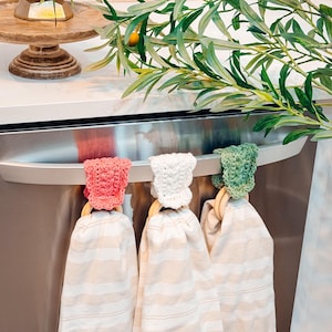 May include: Three kitchen towels with striped patterns hang from a stainless steel appliance. Each towel is held by a wooden ring and topped with a crocheted cap in pink, white, and green. Olive branches and a wooden cake stand are in the background.