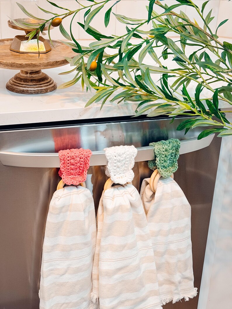 May include: Three kitchen towels with striped patterns hang from a stainless steel appliance. Each towel is held by a wooden ring and topped with a crocheted cap in pink, white, and green. Olive branches and a wooden cake stand are in the background.