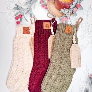 May include: Three crocheted stockings in cream, burgundy, and olive green. Each stocking features a textured cable knit pattern, a tan leather patch with a logo, and a wooden tag attached with a jute string and three small wooden beads.