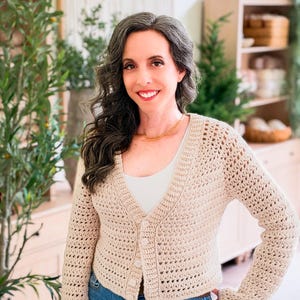 May include: A beige long-sleeved crochet cardigan featuring a V-neckline, a button-down front with four matching buttons, and a textured open-stitch pattern. The garment is worn over a plain white top and paired with blue denim jeans.