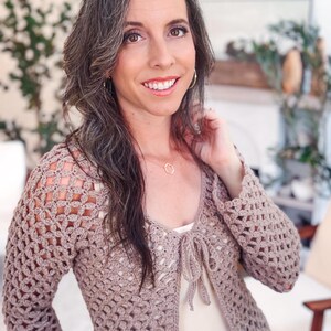 May include: A long-sleeved, taupe-colored crochet cardigan featuring an open-work, repeating geometric pattern throughout the body and sleeves. The garment has a V-neckline and a front tie closure made of matching yarn, worn over a light-colored top.