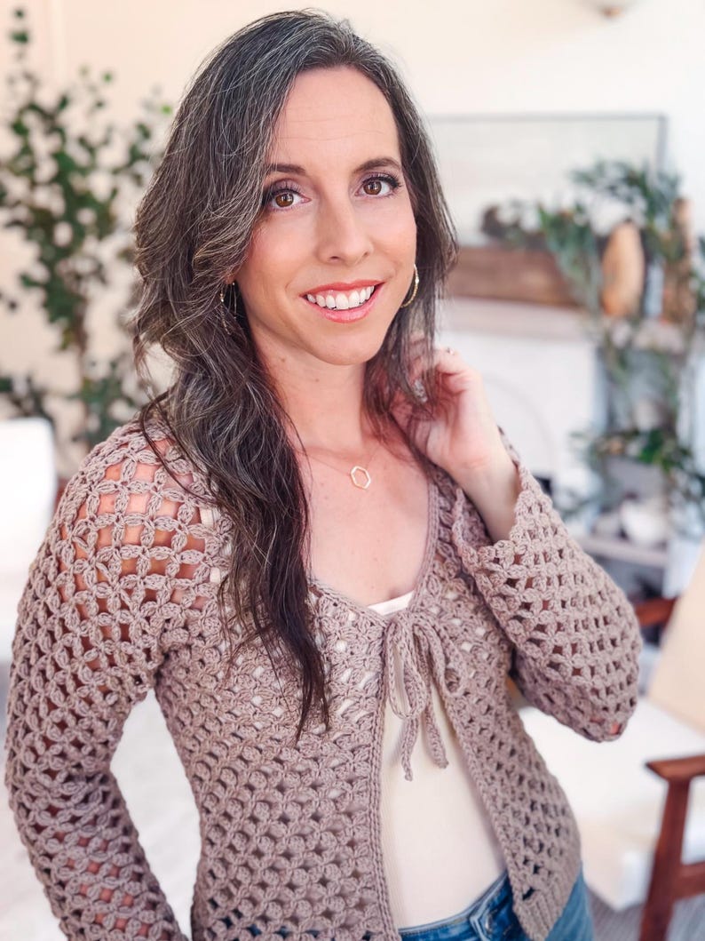 May include: A long-sleeved, taupe-colored crochet cardigan featuring an open-work, repeating geometric pattern throughout the body and sleeves. The garment has a V-neckline and a front tie closure made of matching yarn, worn over a light-colored top.