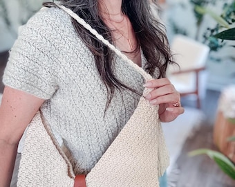 May include: A cream-colored crocheted shoulder bag featuring a braided strap and a brown leather tab closure with a silver-toned metal snap button. The bag has a textured, woven appearance and is worn over the shoulder with a light-colored knit top.
