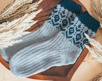 May include: A pair of gray and teal knitted socks with a geometric pattern. The socks are lying on a wooden surface with dried pampas grass.