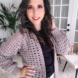 A smiling woman with curly hair wearing a beige crochet cardigan, black top, and white pants, standing indoors, accessorized with unique jewellery.