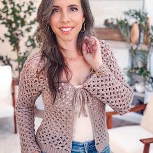 May include: A light brown, long-sleeved crochet cardigan featuring an open-work floral pattern, a tie-front closure, and a solid ribbed hem. The garment is worn over a plain cream-colored tank top and blue denim jeans.