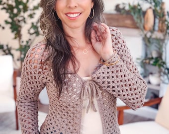May include: A light brown, long-sleeved crochet cardigan featuring an open-work floral pattern, a tie-front closure, and a solid ribbed hem. The garment is worn over a plain cream-colored tank top and blue denim jeans.