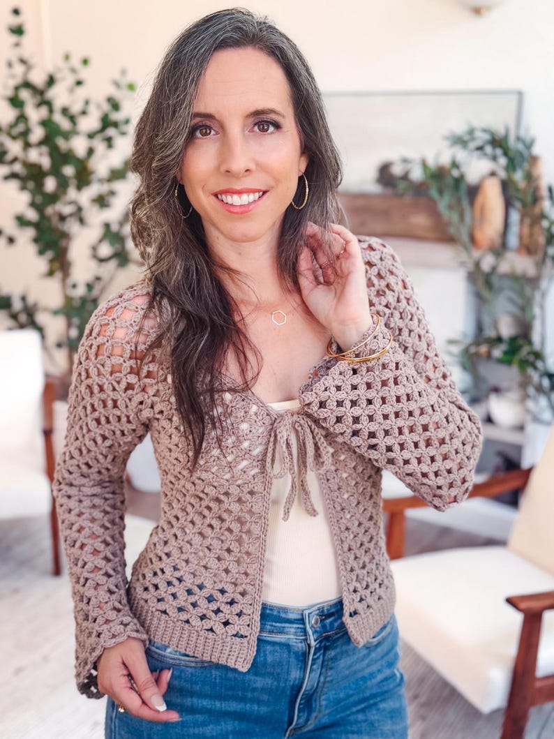 May include: A light brown, long-sleeved crochet cardigan featuring an open-work floral pattern, a tie-front closure, and a solid ribbed hem. The garment is worn over a plain cream-colored tank top and blue denim jeans.