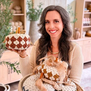 May include: A collection of handmade crochet pumpkins featuring a granny square pattern in cream, brown, dark blue, and olive green. Each pumpkin is topped with a cinnamon stick stem and a burlap ribbon bow, creating a textured, soft appearance.