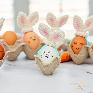 May include: A carton of decorated Easter eggs. Some eggs are brown, others are white with bunny faces and crochet bunny ears. The eggs are in a cardboard egg carton. The bunny ears are white with pink inner ears.