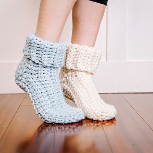 May include: A pair of crocheted slipper socks, one in light blue and one in white, with a folded cuff. The socks are worn on a person's feet, standing on a wooden floor.