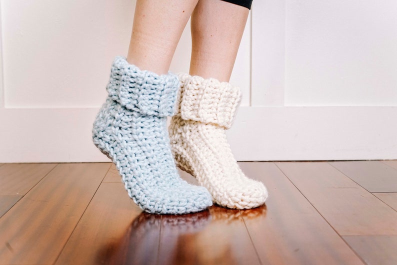 May include: A pair of crocheted slipper socks, one in light blue and one in white, with a folded cuff. The socks are worn on a person's feet, standing on a wooden floor.