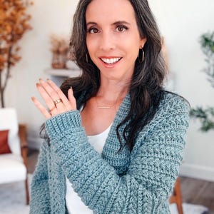 May include: A light blue crochet cardigan with a textured stitch pattern. The cardigan has long sleeves and a relaxed fit. The person is wearing a gold ring and gold hoop earrings. The cardigan is a handmade item.