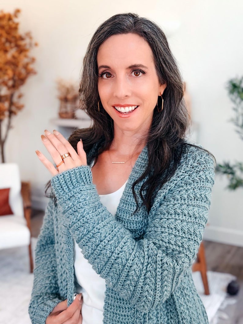 May include: A light blue crochet cardigan with a textured stitch pattern. The cardigan has long sleeves and a relaxed fit. The person is wearing a gold ring and gold hoop earrings. The cardigan is a handmade item.