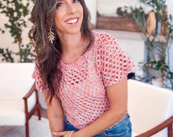 Op de afbeelding: Een roze gehaakte top met korte mouwen en een ronde halslijn. De top wordt gecombineerd met een blauwe jeans. De vrouw draagt gouden oorbellen en armbanden. De achtergrond omvat witte stoelen en groen.