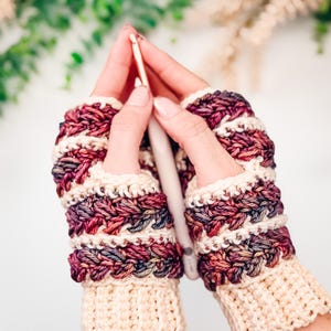 May include: Crocheted fingerless gloves in cream, burgundy, and purple. The gloves have a textured stitch pattern and a cream cuff. A crochet hook is held between the gloves, suggesting a crafting theme.