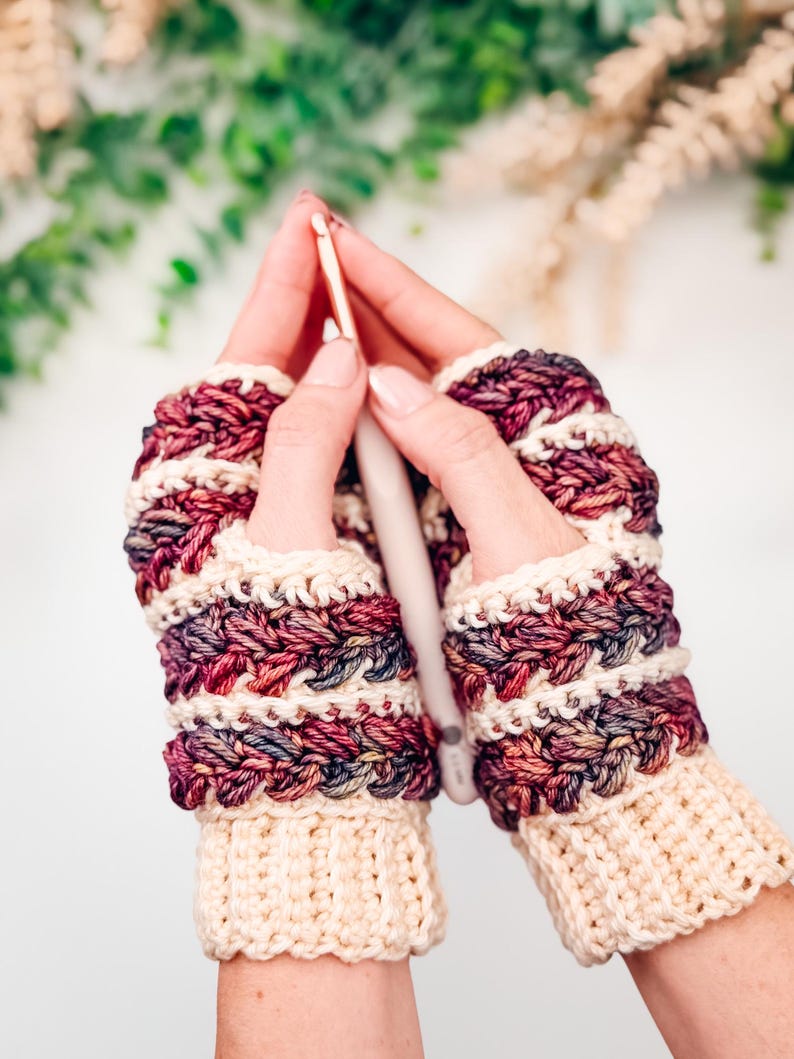 May include: Crocheted fingerless gloves in cream, burgundy, and purple. The gloves have a textured stitch pattern and a cream cuff. A crochet hook is held between the gloves, suggesting a crafting theme.
