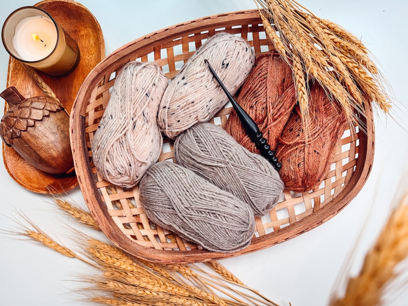 A woven basket holds four skeins of yarn in various shades, a crochet hook, a candle, and wheat stalks on a light surface—perfect for creating a handmade gift.