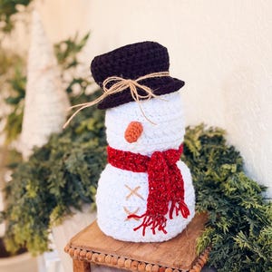 White crochet snowman with brown top hat and sparkly red scarf on a wooden riser surrounded by greenery