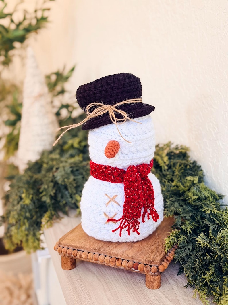 White crochet snowman with brown top hat and sparkly red scarf on a wooden riser surrounded by greenery