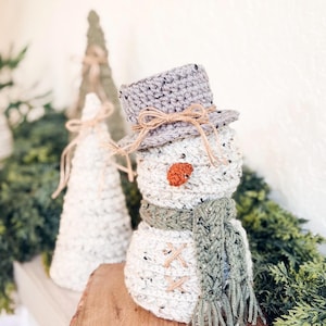 Speckled cream crochet snowman with green scarf beside crochet cone trees on a shelf with greenery