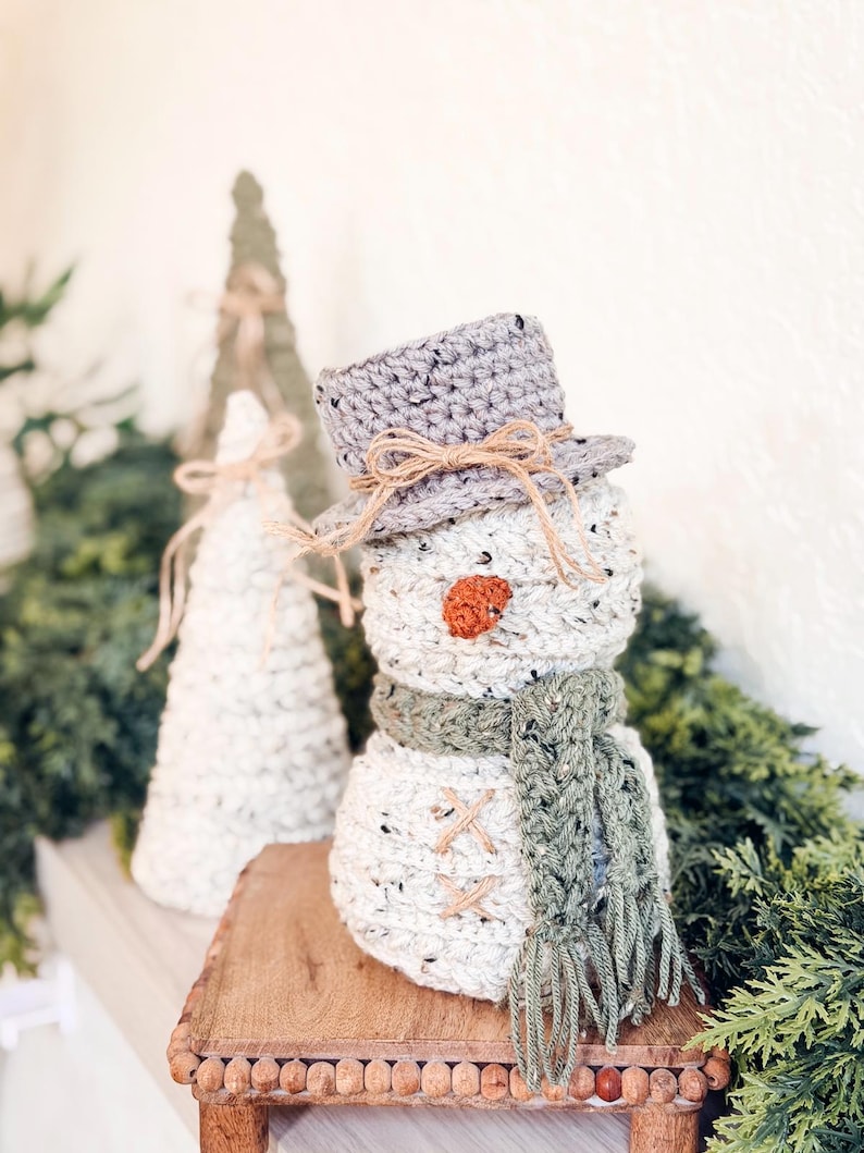 Speckled cream crochet snowman with green scarf beside crochet cone trees on a shelf with greenery