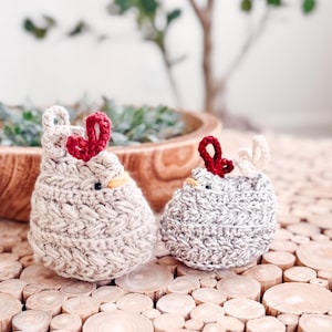 Two small crochet chickens in oatmeal and light gray with red combs sitting side by side on wood slices