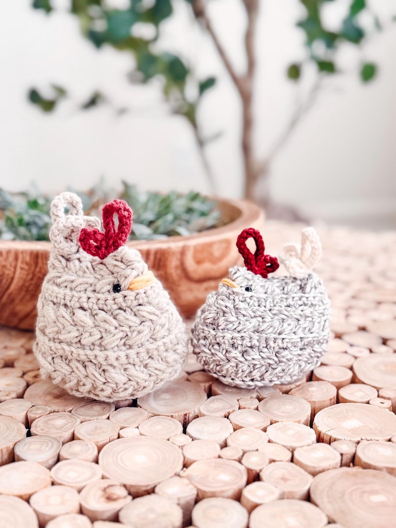 Two small crochet chickens in oatmeal and light gray with red combs sitting side by side on wood slices