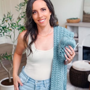 May include: A light blue crocheted shawl with a matching ball of yarn and a crochet hook. The shawl has a textured pattern. The yarn is held in the hand.