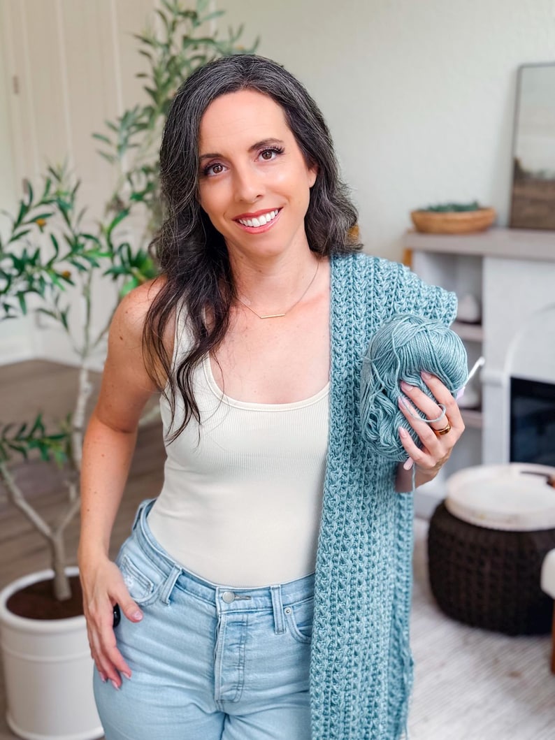 May include: A light blue crocheted shawl with a matching ball of yarn and a crochet hook. The shawl has a textured pattern. The yarn is held in the hand.