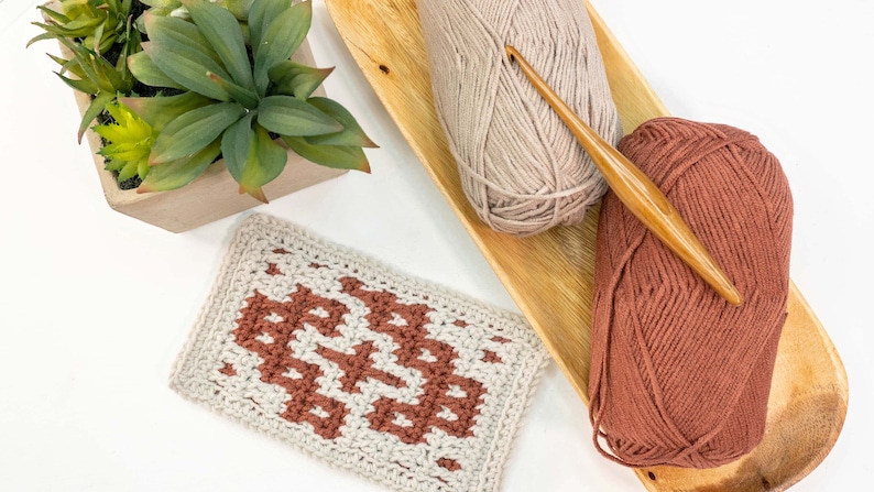 Puede incluir: Un pa&ntilde;o de cocina tejido a crochet con un patr&oacute;n geom&eacute;trico en tonos marrones y beige. Se muestran dos ovillos de lana, uno beige y otro marr&oacute;n, con un gancho de crochet de madera.