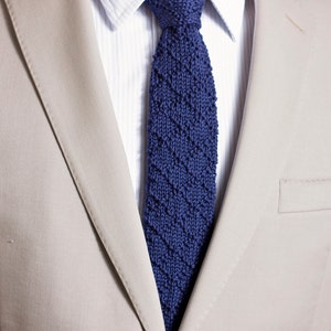 May include: A beige suit jacket with a blue knitted tie. The tie has a diagonal pattern.