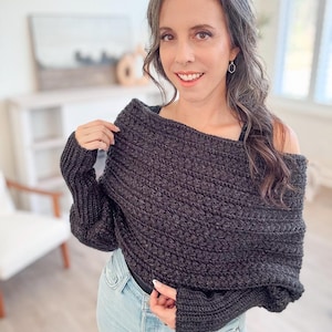 A woman stands indoors wearing a textured black  crochet sweater scarf and light jeans, smiling slightly while holding one side of the sweater. Her unique jewelry gleams subtly against the crochted fabric.