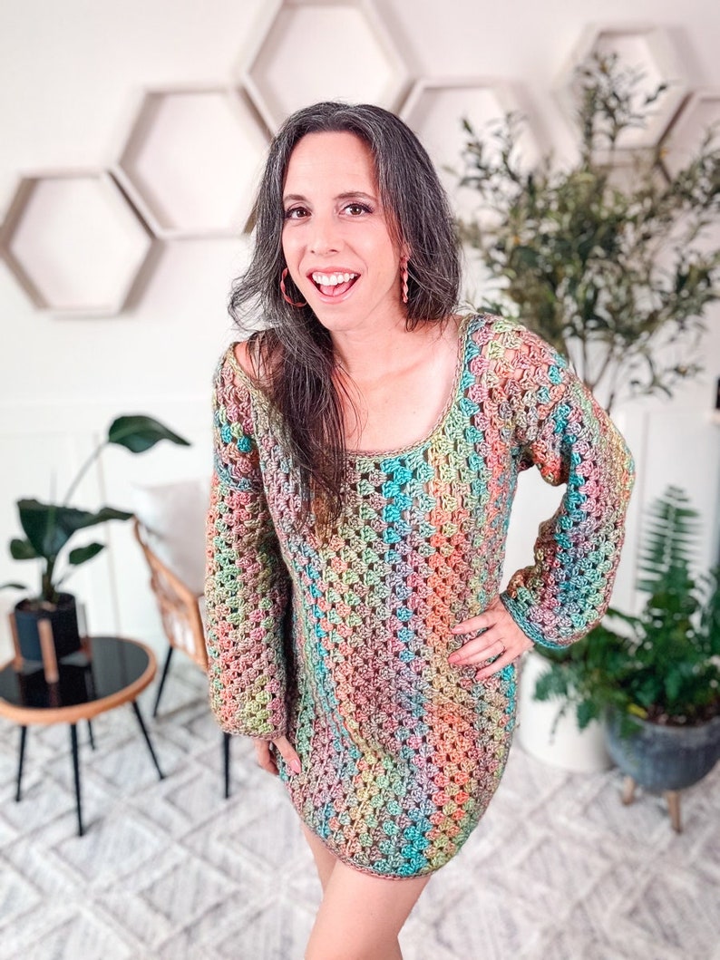 A woman stands indoors wearing a colorful, crocheted dress and unique jewelry. She is smiling, with one hand on her hip. The background features plants and hexagonal wall shelves.