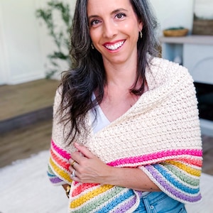 May include: A cream-colored crochet shawl featuring a textured stitch pattern with a vibrant border of horizontal stripes in hot pink, orange, yellow, light green, blue, and purple. The shawl is draped over the shoulders and across the upper body.