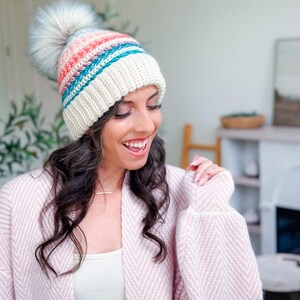 A woman smiles while wearing a crochet hat with colorful stripes and a pom-pom, paired with a textured, light pink cardigan and unique jewellery in a bright indoor setting.