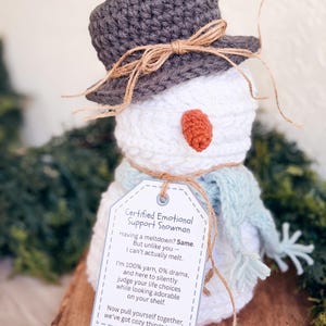 White crochet snowman with gray hat, blue scarf and printed emotional support snowman tag on a wooden riser