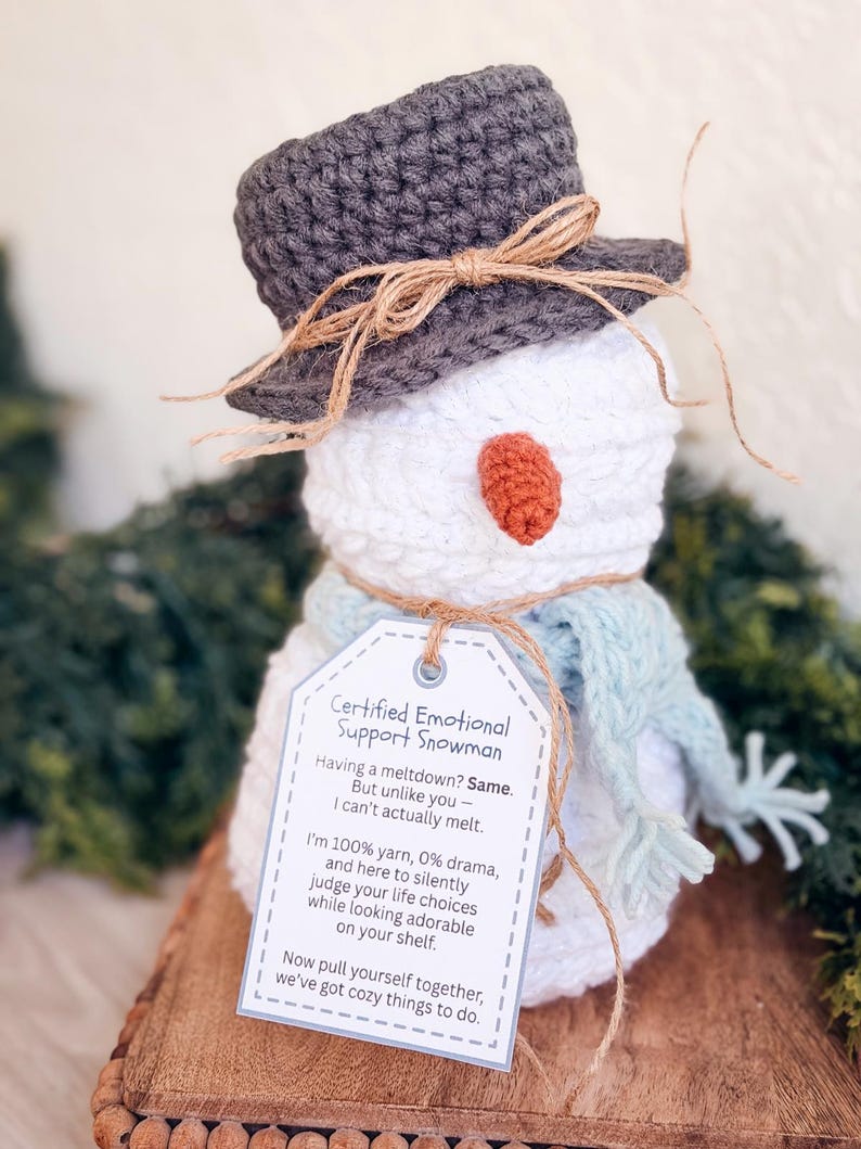 White crochet snowman with gray hat, blue scarf and printed emotional support snowman tag on a wooden riser