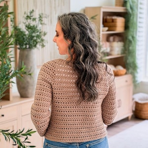 May include: A tan long-sleeved crochet sweater with a textured, open-stitch pattern and a round neckline, paired with blue denim jeans. The sweater features a consistent horizontal weave throughout the body and sleeves, ending at the waistline.