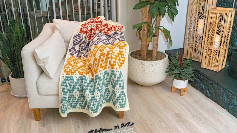 Puede incluir: Un sill&oacute;n blanco con una manta de ganchillo de colores drapeada sobre &eacute;l. La manta tiene rayas de naranja, morado, azul verdoso y rojo.
