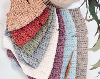 May include: A collection of stacked crochet sweaters in various solid colors including beige, rust, sage green, white, plum, and light blue. Each sweater features a textured cable knit pattern and a high neckline. A wooden crochet hook is placed on top.