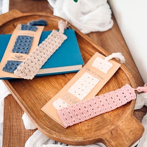 Puede incluir: Cuatro marcapáginas rectangulares de ganchillo en azul, beige, blanco y rosa, cada uno con un patrón calado texturizado y una pequeña borla en la parte superior. Dos marcapáginas están sujetos a tarjetas de papel beige, una con el texto one of a kind.