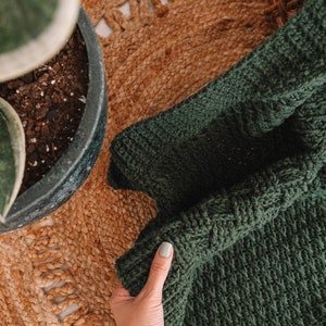 Puede incluir: Una manta de ganchillo de color verde bosque oscuro con un patr&oacute;n de punto de cester&iacute;a denso y texturizado. La tela es gruesa y uniforme, mostrando una construcci&oacute;n de hilo consistente en toda la pieza, destacando los intrincados bucles entrelazados.